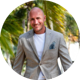 Leigh in a light grey blazer and white shirt stands outdoors in front of palm trees, smiling at the camera.