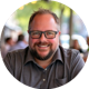 Andy with glasses and a beard smiles while seated outdoors at a table, with blurred people and trees in the background.